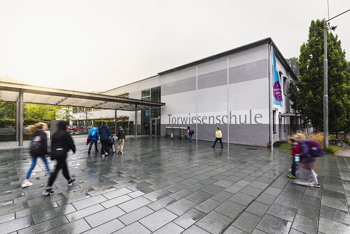 Der Haupteingang und das vordere Gebäude der Torwiesenschule an einem verregneten Morgen. Schüler bewegen sich Richtung Eingang.